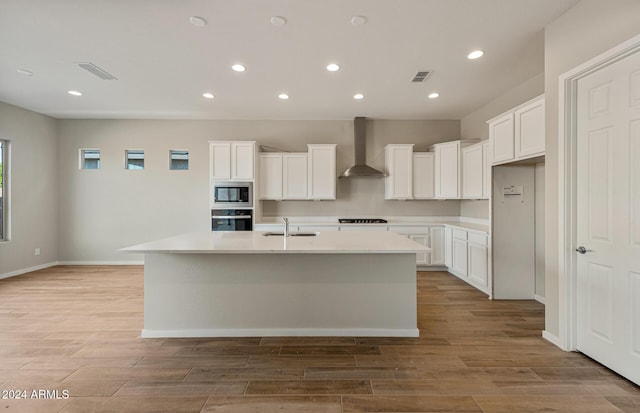 kitchen with wall chimney exhaust hood, stainless steel oven, built in microwave, a center island with sink, and white cabinets