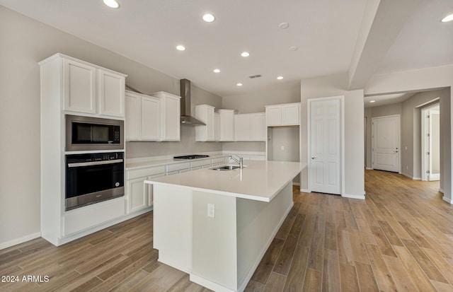 kitchen featuring built in microwave, wall chimney exhaust hood, oven, a center island with sink, and white cabinets