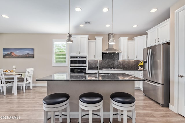 kitchen featuring wall chimney exhaust hood, stainless steel appliances, pendant lighting, a center island with sink, and white cabinets