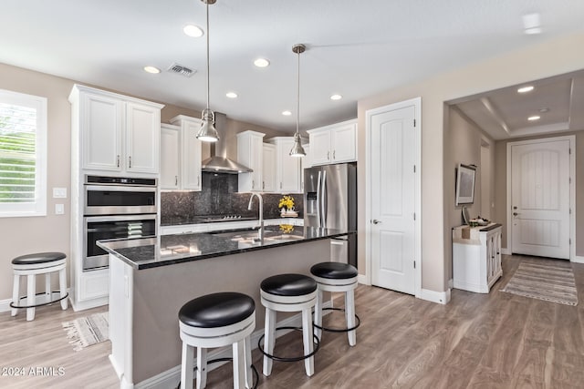 kitchen featuring stainless steel appliances, wall chimney range hood, light hardwood / wood-style floors, white cabinetry, and an island with sink