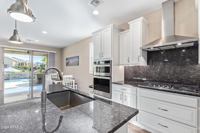 kitchen with appliances with stainless steel finishes, sink, wall chimney range hood, dark stone countertops, and white cabinets