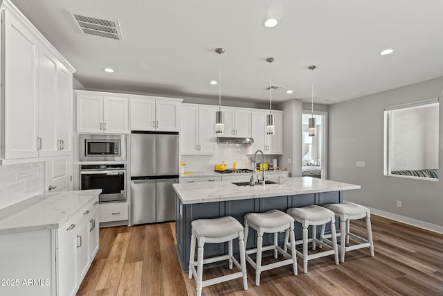 kitchen with appliances with stainless steel finishes, white cabinetry, an island with sink, hanging light fixtures, and light stone counters