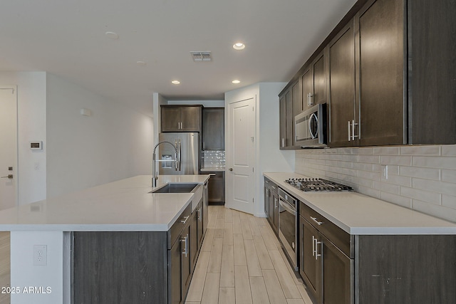 kitchen with stainless steel appliances, a sink, backsplash, dark brown cabinets, and light wood-style floors
