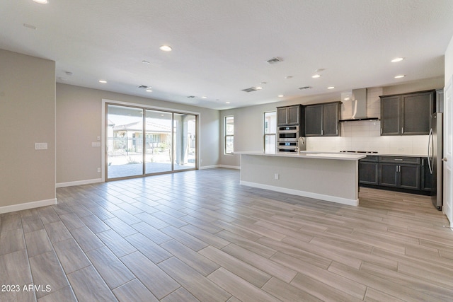 kitchen with wall chimney range hood, a kitchen island with sink, a healthy amount of sunlight, and appliances with stainless steel finishes