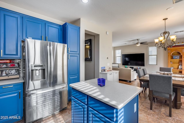 kitchen with stainless steel refrigerator with ice dispenser, ceiling fan with notable chandelier, blue cabinets, pendant lighting, and a center island