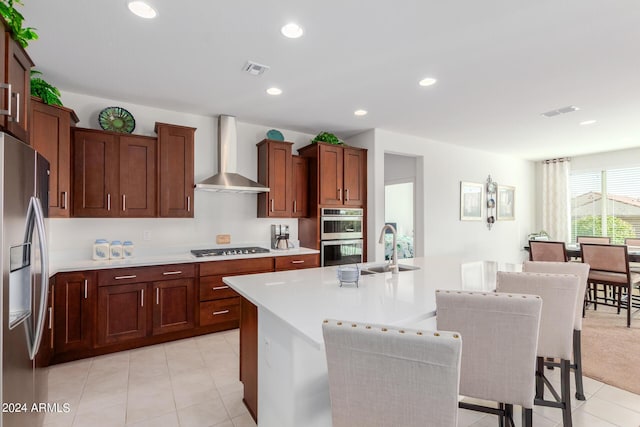 kitchen featuring sink, wall chimney range hood, light tile patterned floors, stainless steel appliances, and a center island with sink