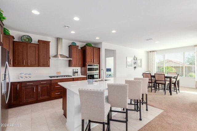 kitchen with appliances with stainless steel finishes, an island with sink, a kitchen bar, light carpet, and wall chimney range hood