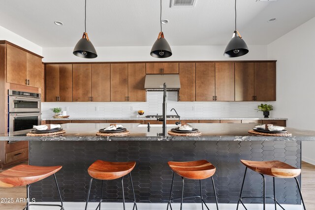 kitchen with double oven, a breakfast bar area, ventilation hood, tasteful backsplash, and decorative light fixtures