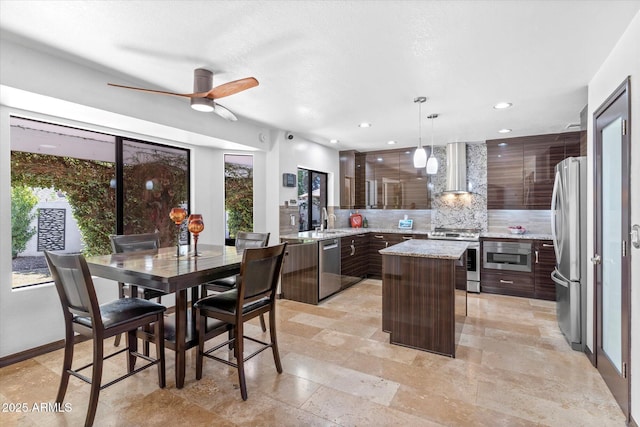 kitchen featuring appliances with stainless steel finishes, hanging light fixtures, a center island, dark brown cabinetry, and wall chimney exhaust hood