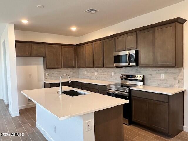 kitchen with appliances with stainless steel finishes, an island with sink, sink, backsplash, and dark brown cabinets