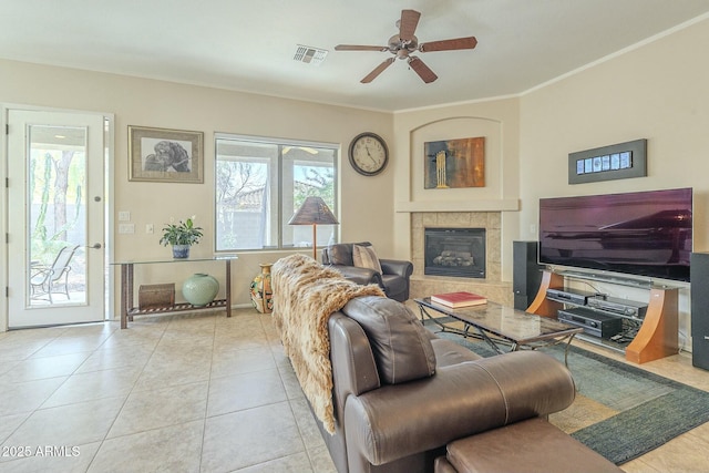 tiled living room with a tiled fireplace, a ceiling fan, visible vents, and ornamental molding