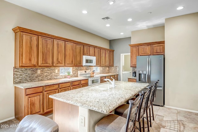 kitchen featuring white appliances, visible vents, brown cabinetry, an island with sink, and backsplash