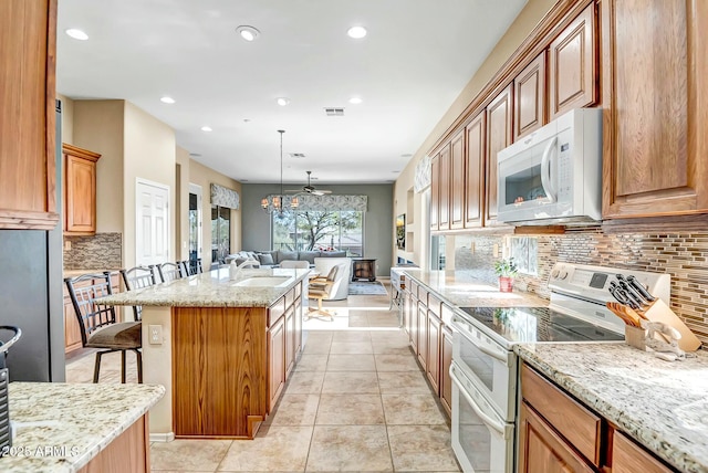 kitchen with light stone counters, white appliances, a kitchen bar, a center island with sink, and pendant lighting