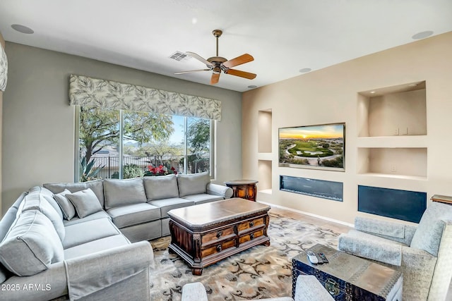 living area with built in shelves, a glass covered fireplace, visible vents, and ceiling fan