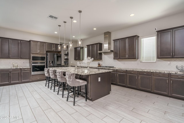 kitchen with appliances with stainless steel finishes, pendant lighting, a kitchen breakfast bar, a kitchen island with sink, and wall chimney range hood