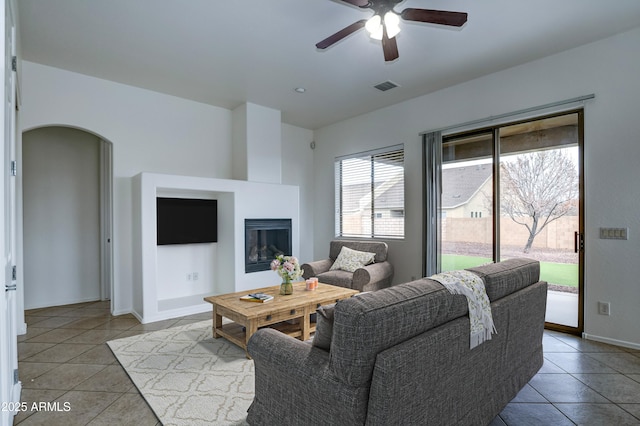 tiled living room with visible vents, a ceiling fan, a glass covered fireplace, arched walkways, and baseboards