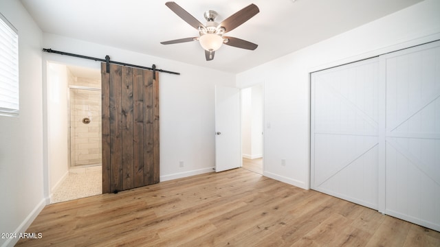 unfurnished bedroom featuring ensuite bath, light hardwood / wood-style flooring, ceiling fan, a barn door, and a closet