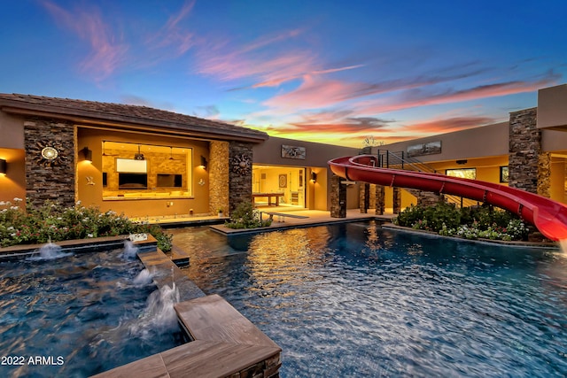 pool at dusk featuring a patio, pool water feature, and a water slide