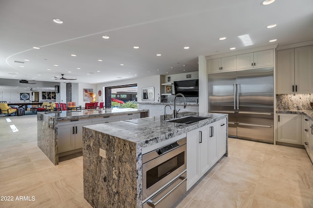 kitchen featuring ceiling fan, dark stone countertops, backsplash, a center island with sink, and stainless steel appliances