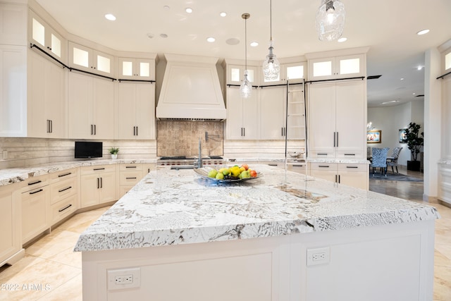 kitchen featuring a kitchen island with sink, light stone counters, custom exhaust hood, tasteful backsplash, and decorative light fixtures