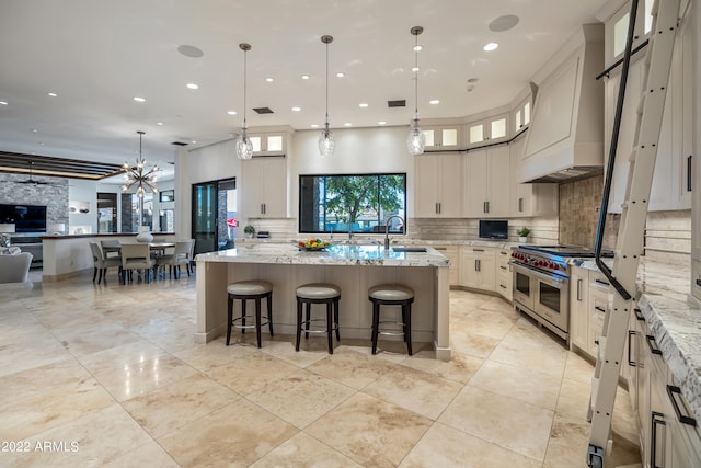 kitchen with decorative light fixtures, custom exhaust hood, an island with sink, and light stone counters