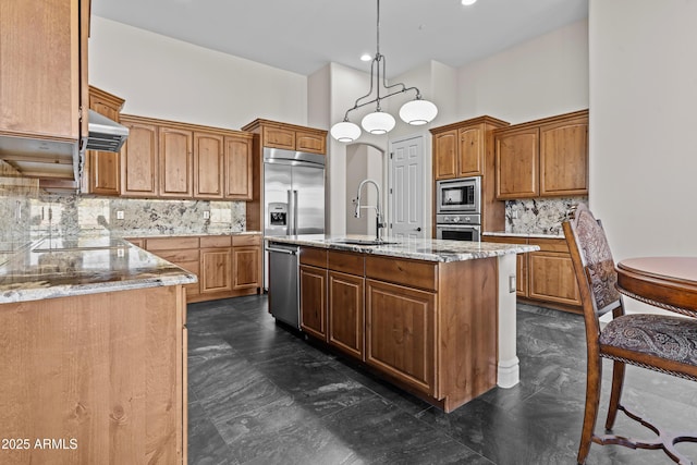 kitchen with decorative light fixtures, sink, a kitchen island with sink, built in appliances, and wall chimney range hood