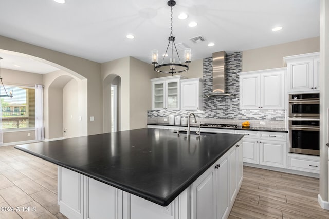 kitchen with white cabinets, a kitchen island with sink, wall chimney range hood, appliances with stainless steel finishes, and an inviting chandelier