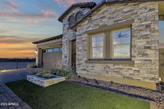 property exterior at dusk featuring a garage, stone siding, driveway, and fence