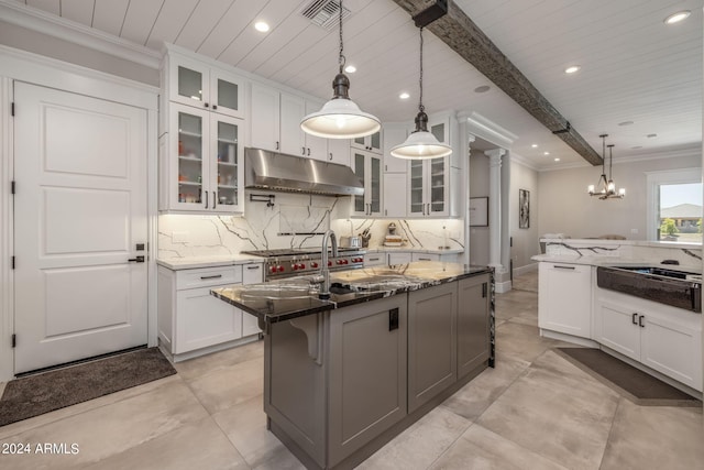 kitchen featuring beam ceiling, decorative light fixtures, dark stone countertops, white cabinets, and an island with sink