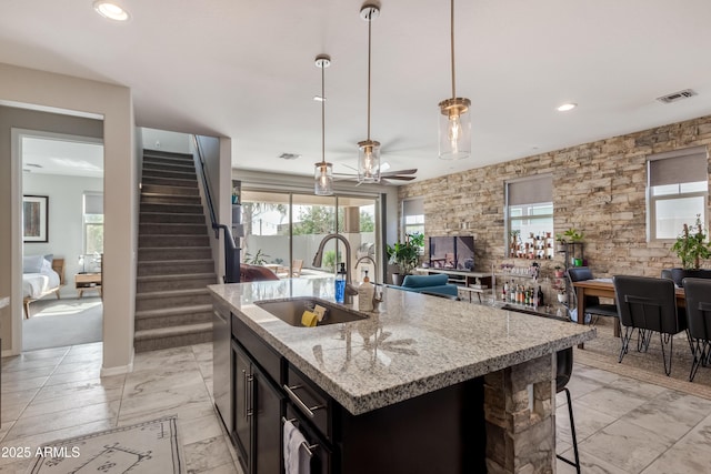 kitchen with pendant lighting, sink, ceiling fan, light stone countertops, and a center island with sink