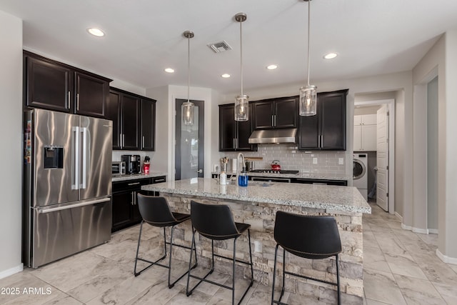 kitchen with pendant lighting, appliances with stainless steel finishes, a kitchen island with sink, light stone countertops, and washer / clothes dryer