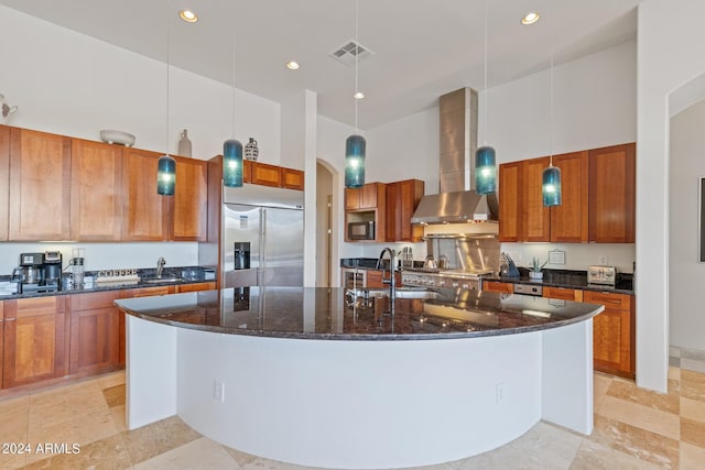 kitchen featuring built in refrigerator, an island with sink, hanging light fixtures, and wall chimney range hood