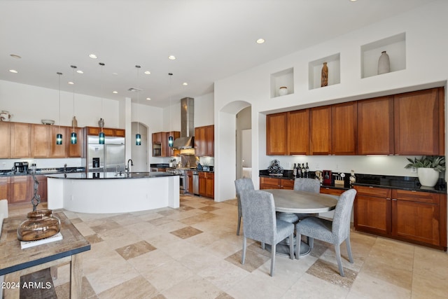 kitchen featuring a towering ceiling, wall chimney exhaust hood, stainless steel appliances, decorative light fixtures, and a center island with sink