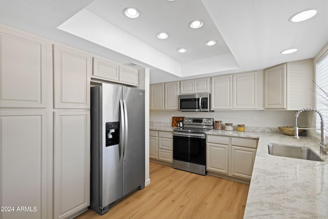 kitchen featuring appliances with stainless steel finishes, light stone counters, a raised ceiling, sink, and light hardwood / wood-style flooring