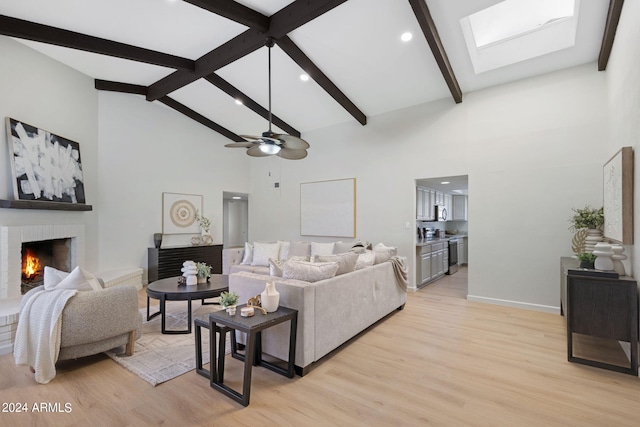 living room with a skylight, a brick fireplace, ceiling fan, beamed ceiling, and light hardwood / wood-style floors