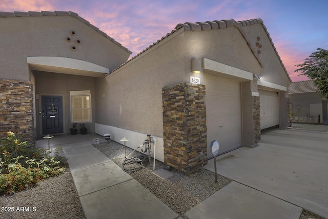 exterior space featuring a tiled roof, stucco siding, stone siding, an attached garage, and concrete driveway