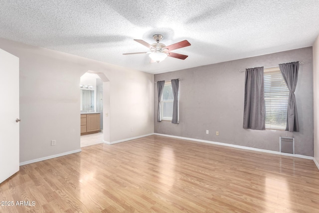 empty room featuring arched walkways, ceiling fan, baseboards, light wood-type flooring, and a textured ceiling