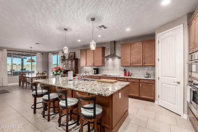 kitchen with wall chimney exhaust hood, black electric stovetop, light stone counters, pendant lighting, and a center island with sink