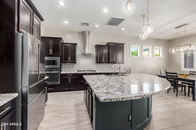 kitchen featuring decorative light fixtures, wall chimney exhaust hood, appliances with stainless steel finishes, a center island with sink, and dark brown cabinetry