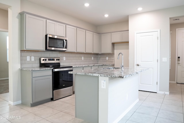 kitchen with sink, light stone counters, an island with sink, stainless steel appliances, and decorative backsplash