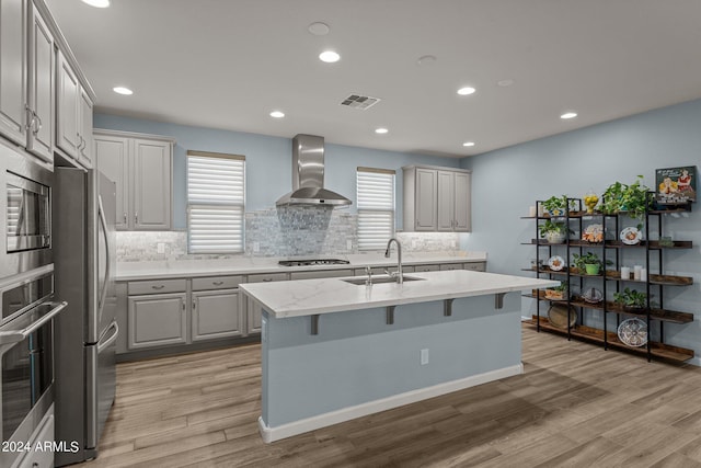 kitchen featuring sink, wall chimney exhaust hood, stainless steel appliances, light hardwood / wood-style floors, and a kitchen island with sink