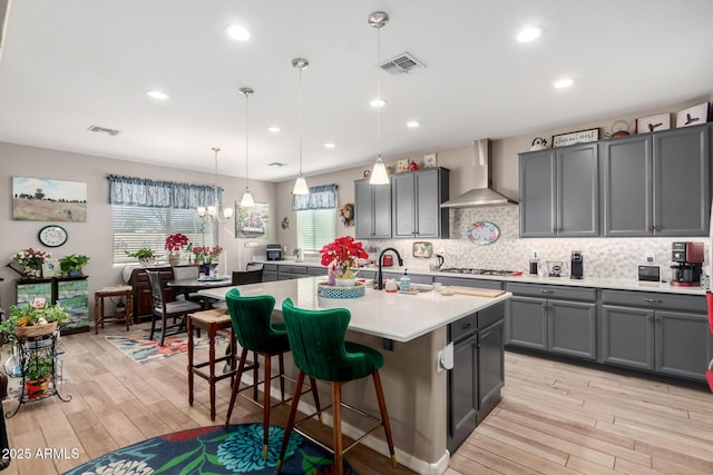 kitchen featuring stainless steel gas cooktop, a kitchen island with sink, decorative light fixtures, wall chimney exhaust hood, and gray cabinets