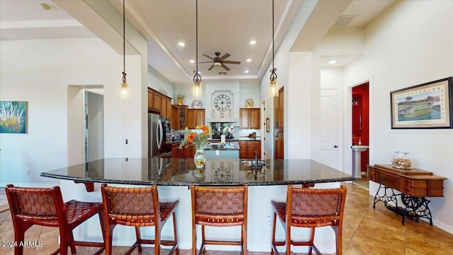 kitchen with ceiling fan, pendant lighting, stainless steel refrigerator, a breakfast bar, and a kitchen island with sink