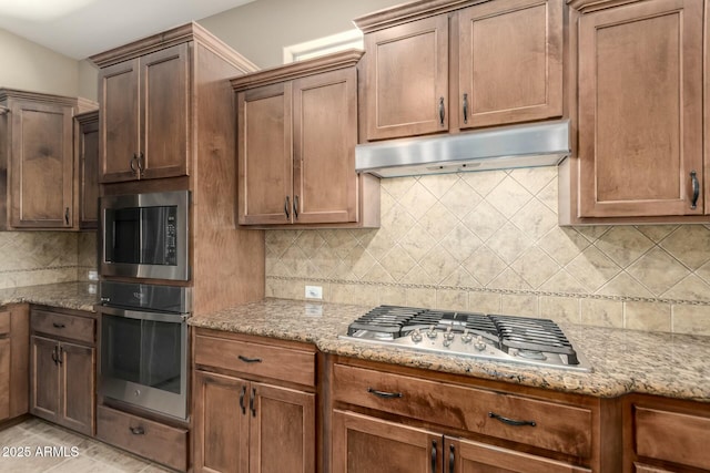 kitchen with stainless steel appliances, under cabinet range hood, decorative backsplash, and light stone countertops