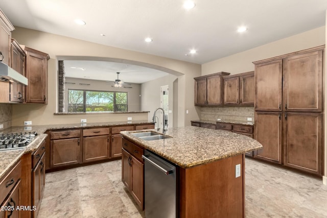 kitchen with a sink, appliances with stainless steel finishes, ceiling fan, light stone countertops, and an island with sink