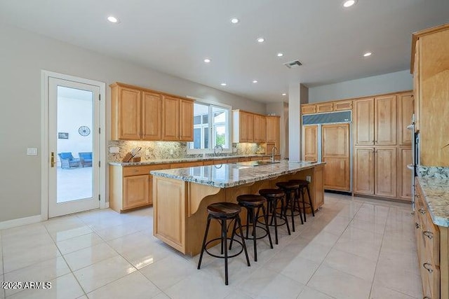 kitchen with light stone counters, visible vents, a center island with sink, paneled refrigerator, and backsplash