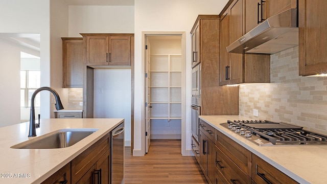 kitchen with wood finished floors, brown cabinetry, a sink, appliances with stainless steel finishes, and under cabinet range hood