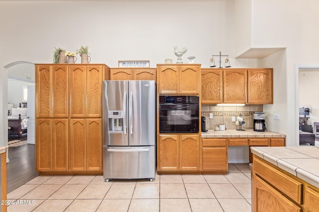 kitchen featuring stainless steel refrigerator with ice dispenser, light tile patterned floors, black oven, and tile counters