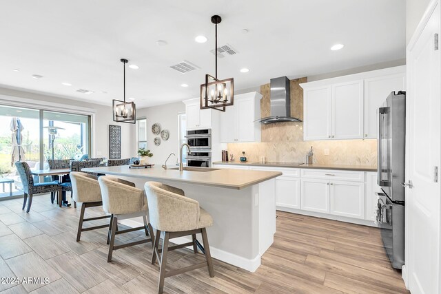 kitchen featuring decorative light fixtures, appliances with stainless steel finishes, a chandelier, wall chimney range hood, and a kitchen island with sink