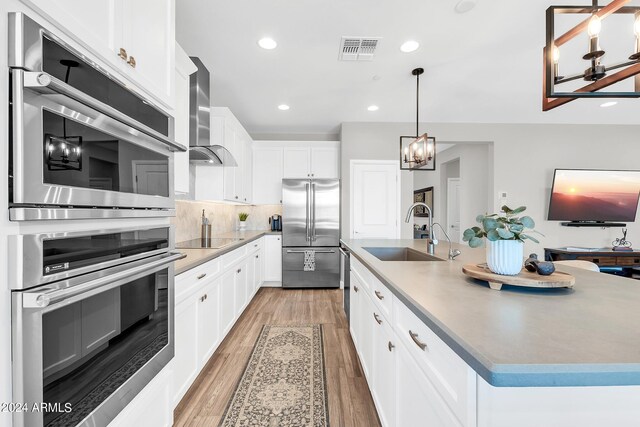 kitchen with wall chimney exhaust hood, appliances with stainless steel finishes, an island with sink, sink, and light wood-type flooring